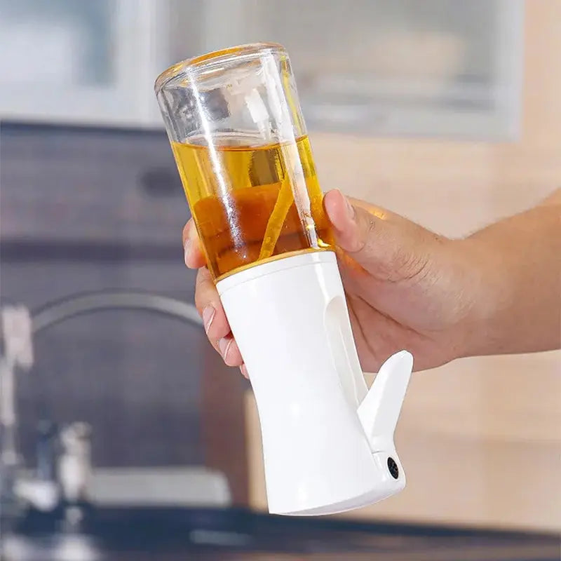 Hand holding a white and clear glass oil dispenser with liquid inside, blurred kitchen background