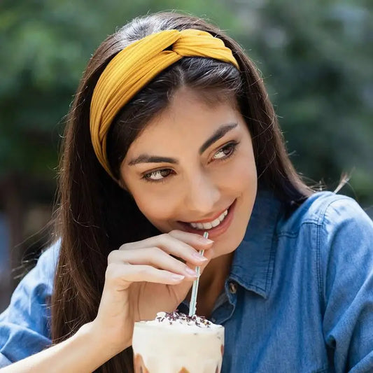 Woman wearing yellow twist headband and blue shirt, drinking from a cup with a straw outdoors – casual summer fashion style