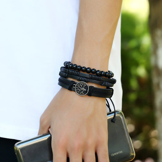 
Hand wearing multiple black braided leather bracelets with Tree of Life charm – outdoor natural background

