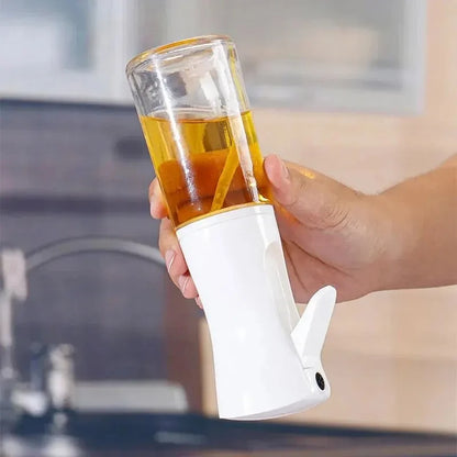 
"Hand holding a white kitchen tool attached to a transparent container filled with oil, designed for controlled pouring or spraying in cooking."